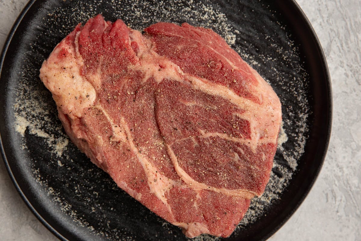 Black plate with a large chuck roast sprinkled with salt, garlic powder, and black pepper.