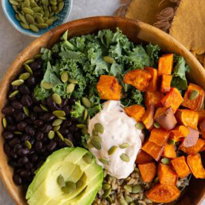 A wooden bowl filled with kale, black beans, roasted sweet potato cubes, avocado slices, rice, and a dollop of creamy sauce, topped with pumpkin seeds. A bowl of seeds and half a lime are in the background.