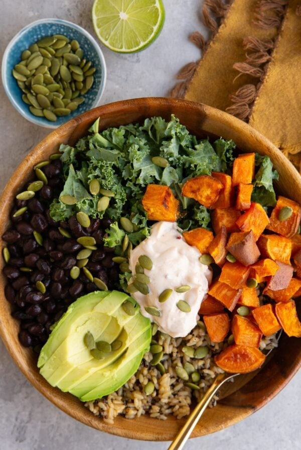A wooden bowl filled with kale, black beans, roasted sweet potato cubes, avocado slices, rice, and a dollop of creamy sauce, topped with pumpkin seeds. A bowl of seeds and half a lime are in the background.