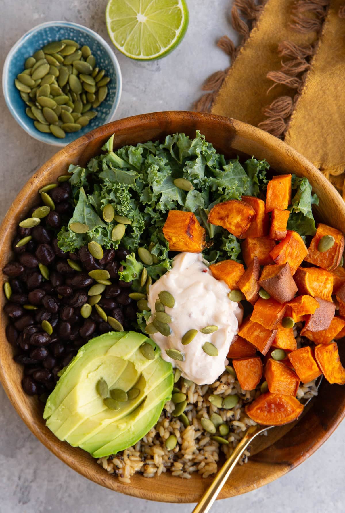 A wooden bowl filled with kale, black beans, roasted sweet potato cubes, avocado slices, rice, and a dollop of creamy sauce, topped with pumpkin seeds. A bowl of seeds and half a lime are in the background.