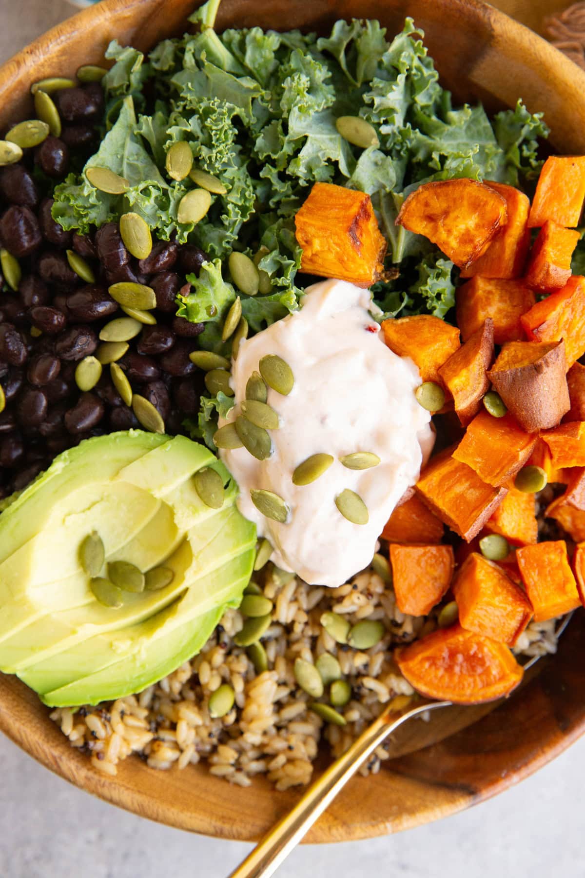 A bowl filled with kale, black beans, roasted sweet potato cubes, sliced avocado, brown rice, pepitas, and a dollop of creamy dressing, arranged in colorful sections.