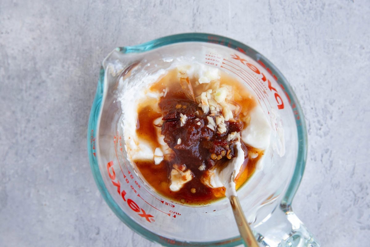 A glass measuring cup containing yogurt, minced garlic, chipotle peppers in adobo sauce, and honey, with a spoon mixing the ingredients on a light gray surface.