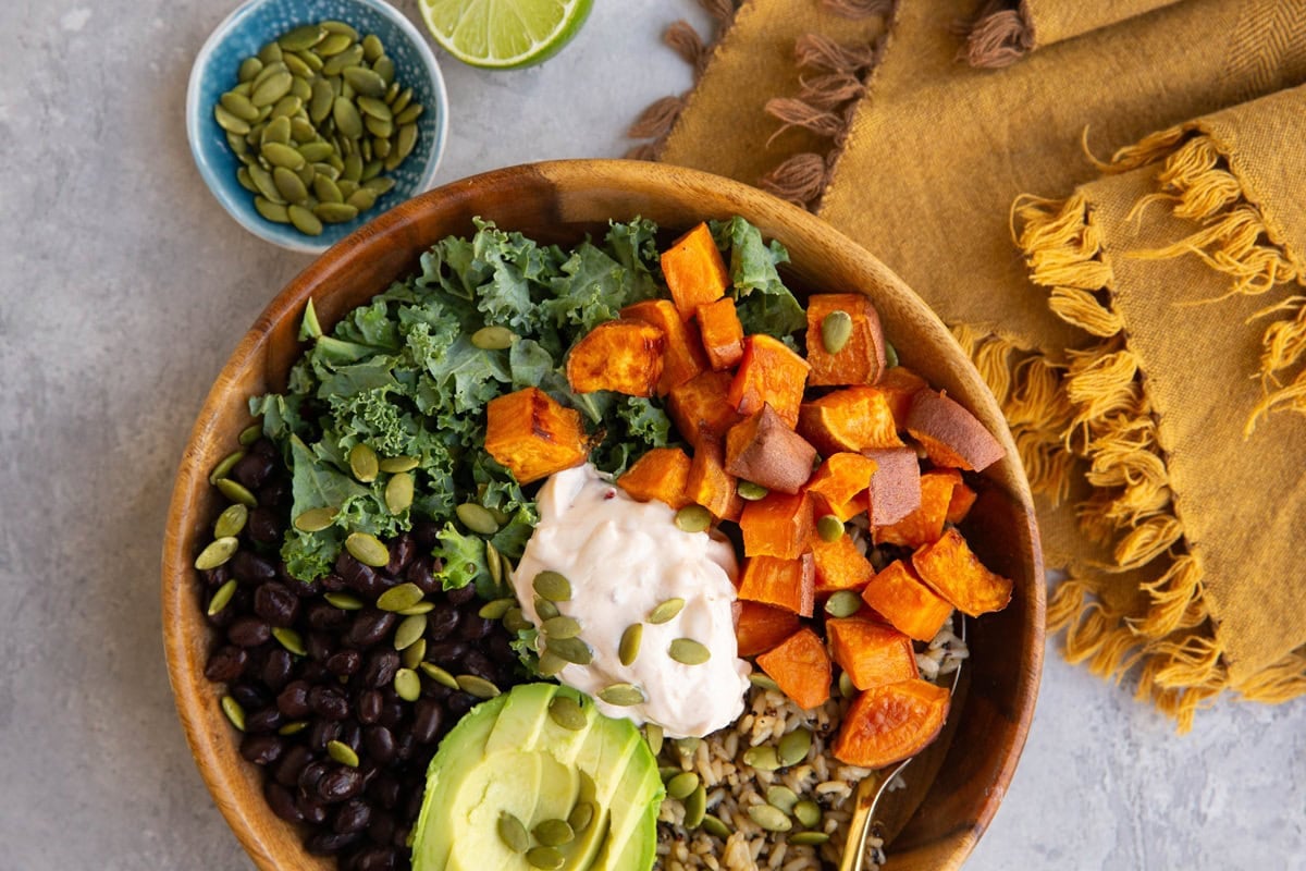 A wooden bowl filled with black beans, kale, roasted sweet potato cubes, avocado slices, wild rice, pumpkin seeds, and a dollop of creamy sauce. A napkin, lime wedge, and small bowl of pumpkin seeds are nearby.