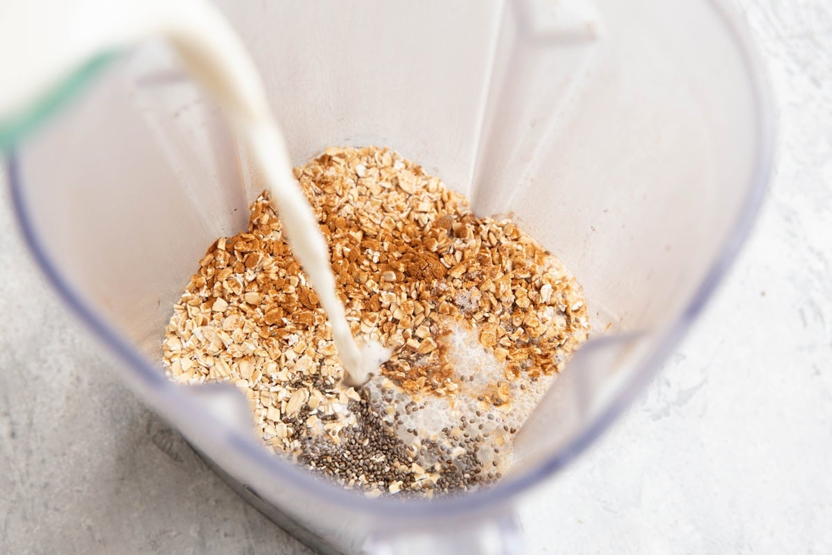 Blender with oats, chia seeds, and cinnamon as milk is being poured in, preparing ingredients for blending.