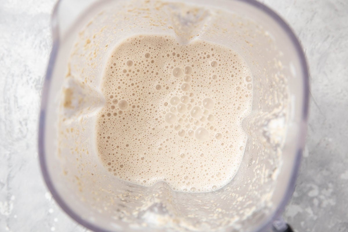 Top-down view of a blender containing a frothy, creamy liquid mixture with bubbles on the surface, set against a light-colored background.