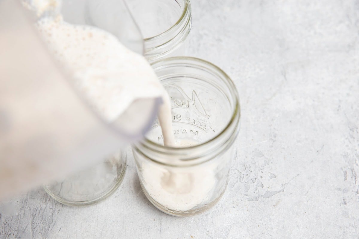 Blended overnight oats being poured from a blender into a clear glass mason jar, with two more empty jars beside it, on a light gray textured surface.