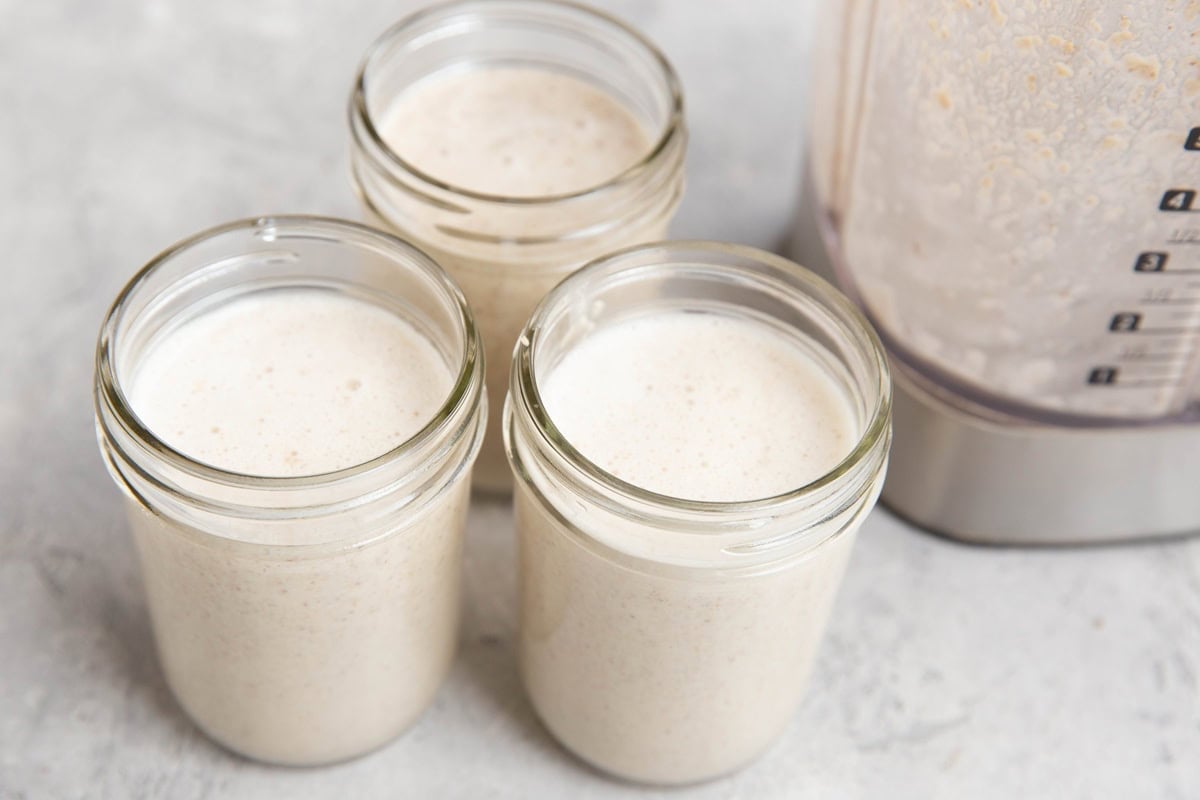 Three glass jars filled with a creamy, light beige smoothie are placed on a light gray surface next to a blender containing more of the same smoothie.