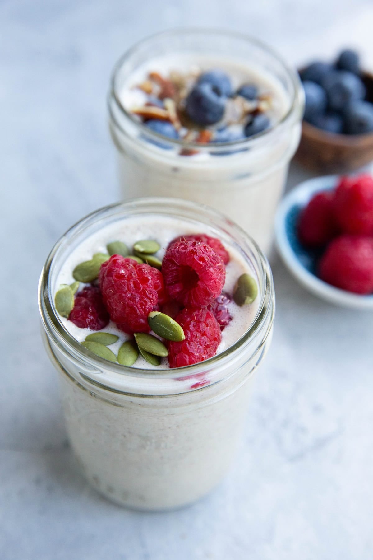 Two jars of overnight oats topped with fresh raspberries and pumpkin seeds, and blueberries with sliced pecans. A small bowl of blueberries and a small plate of raspberries are in the background on a light surface.