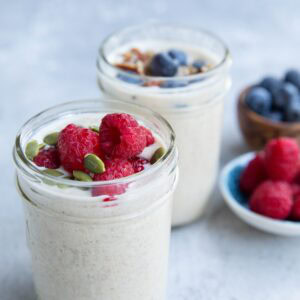 Two glass jars filled with creamy overnight oats, one topped with raspberries and pumpkin seeds, the other with blueberries and nuts. Plates of fresh raspberries and blueberries are in the background on a light surface.
