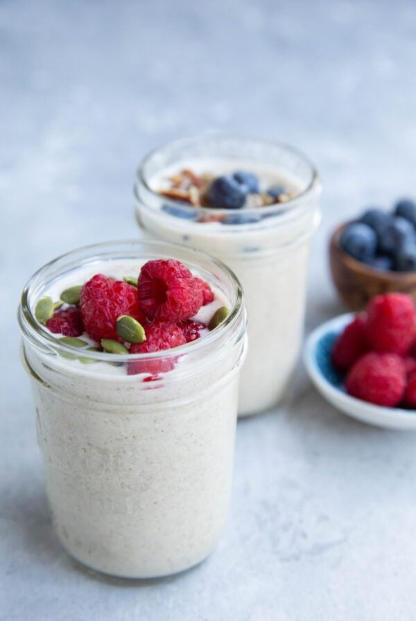 Two glass jars filled with creamy overnight oats, one topped with raspberries and pumpkin seeds, the other with blueberries and nuts. Plates of fresh raspberries and blueberries are in the background on a light surface.
