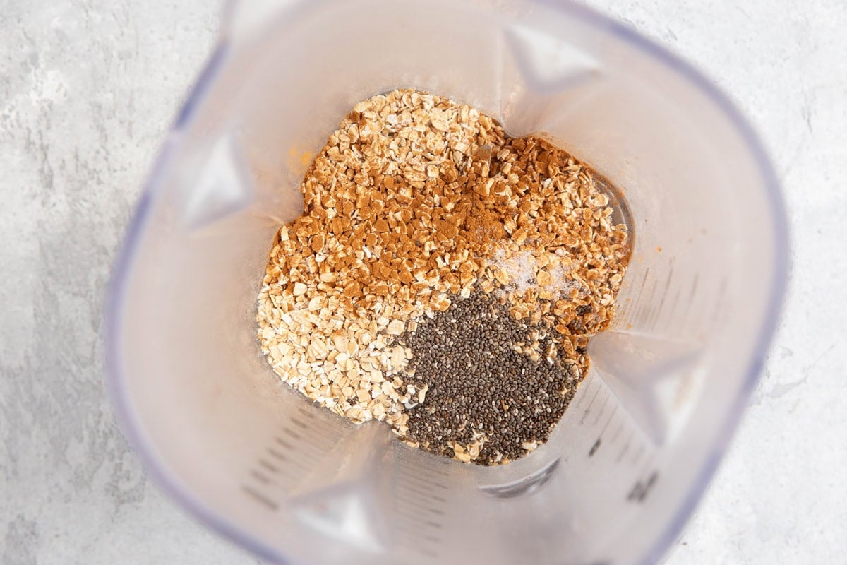 Blender containing dry oats, chia seeds, and ground cinnamon, placed on a light-colored surface.