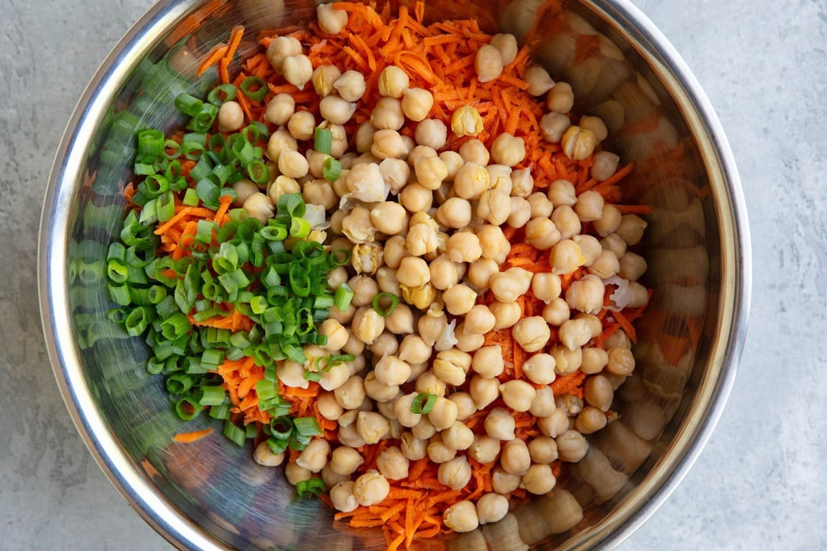 Large stainless steel mixing bowl with grated carrots, chickpeas, and chopped green onions, to be turned into salad.