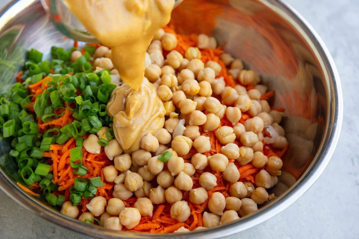 Pouring peanut dressing into a stainless steel mixing bowl full of shredded carrots, chickpeas, and green onions.