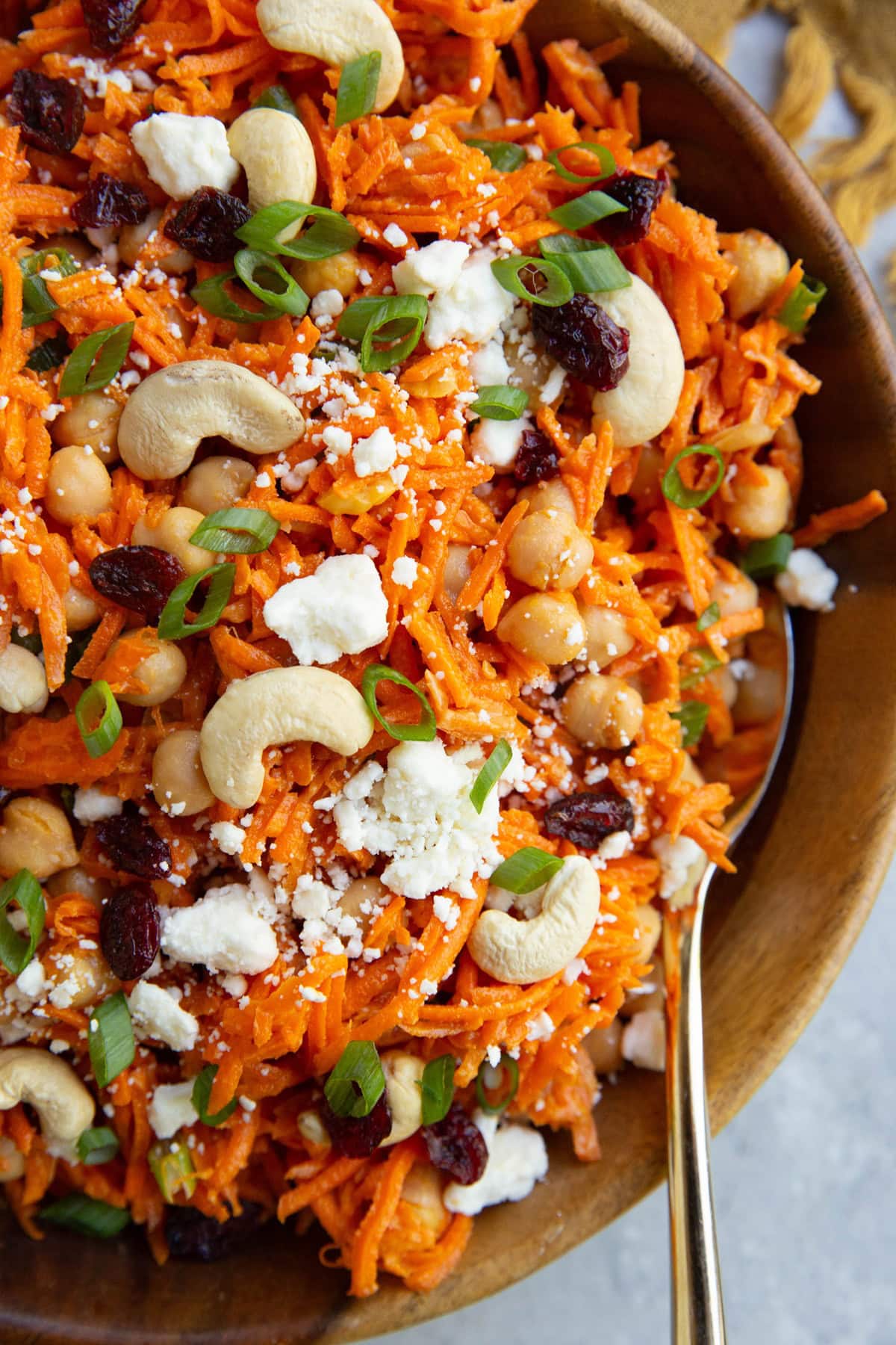 large wooden bowl full of grated carrot salad with green onions, chickpeas, feta cheese, cashews, and dried cranberries.