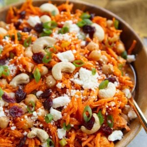Wooden bowl of grated carrots, cashews, dried cranberries, chickpeas, and feta cheese with a golden spoon for serving and a fresh lime in the background.