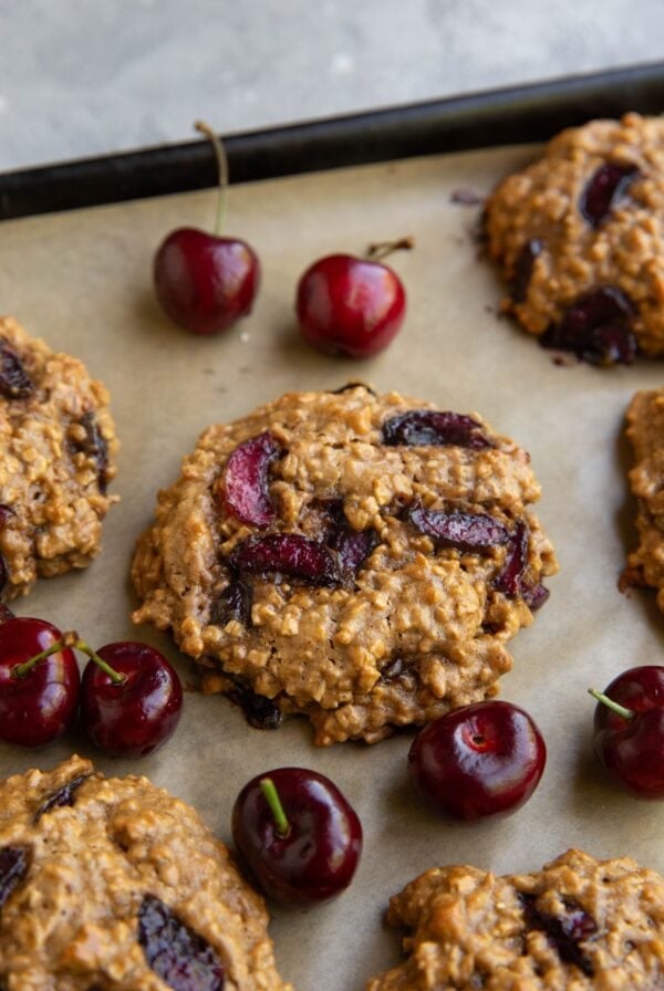 Cherry cookies on a parchment-lined baking sheet with fresh cherries all around.