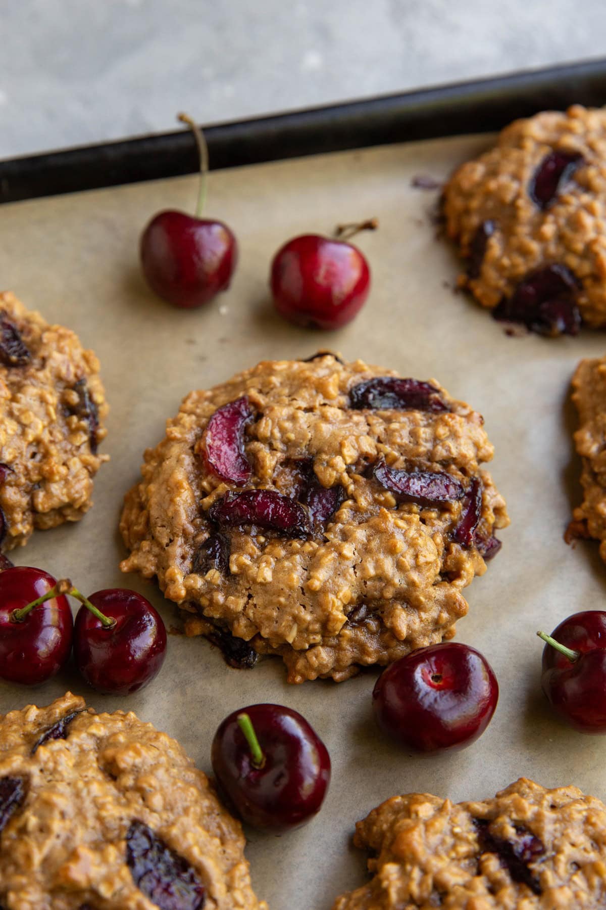 Cherry cookies on a parchment-lined baking sheet with fresh cherries all around.