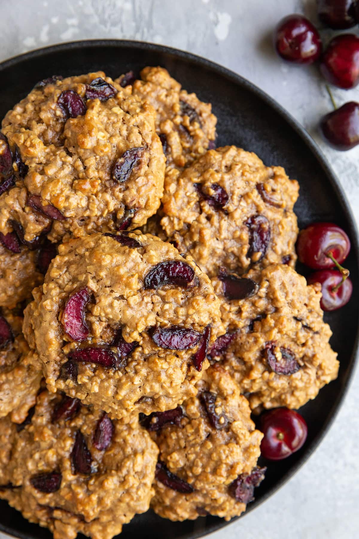 Cherry oatmeal cookies on a black plate with fresh cherries all around.