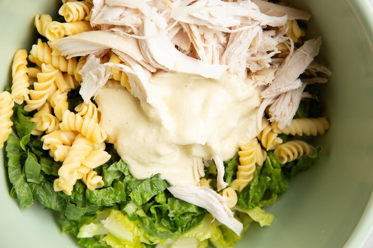 Mixing bowl with chopped lettuce, pasta noodles, chicken, and Caesar dressing, ready to be mixed up.