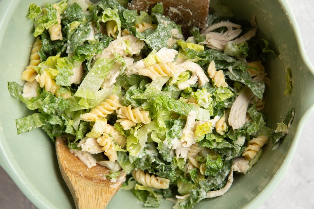 Mixing bowl with chicken Caesar pasta salad mixed up with two wooden spoons.