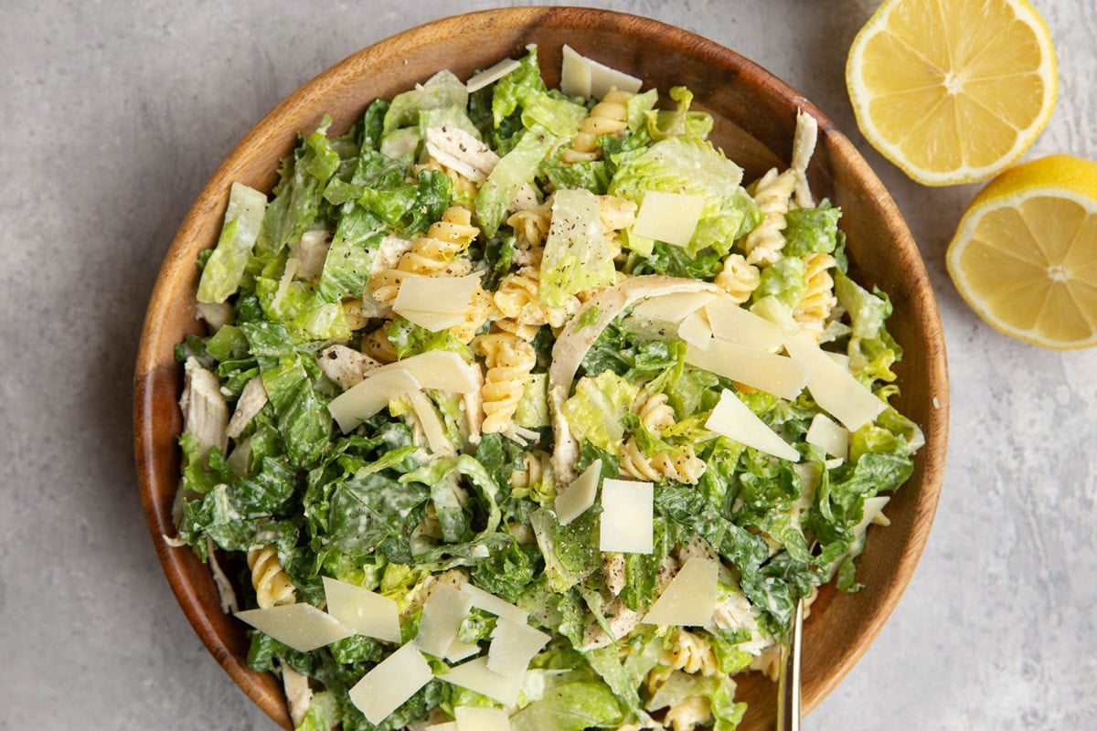 Wooden bowl of chicken Caesar pasta salad with a halved lemon to the side.