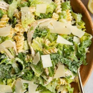Wooden bowl full of chicken Caesar pasta salad with parmesan cheese on top and a sliced lemon to the side.