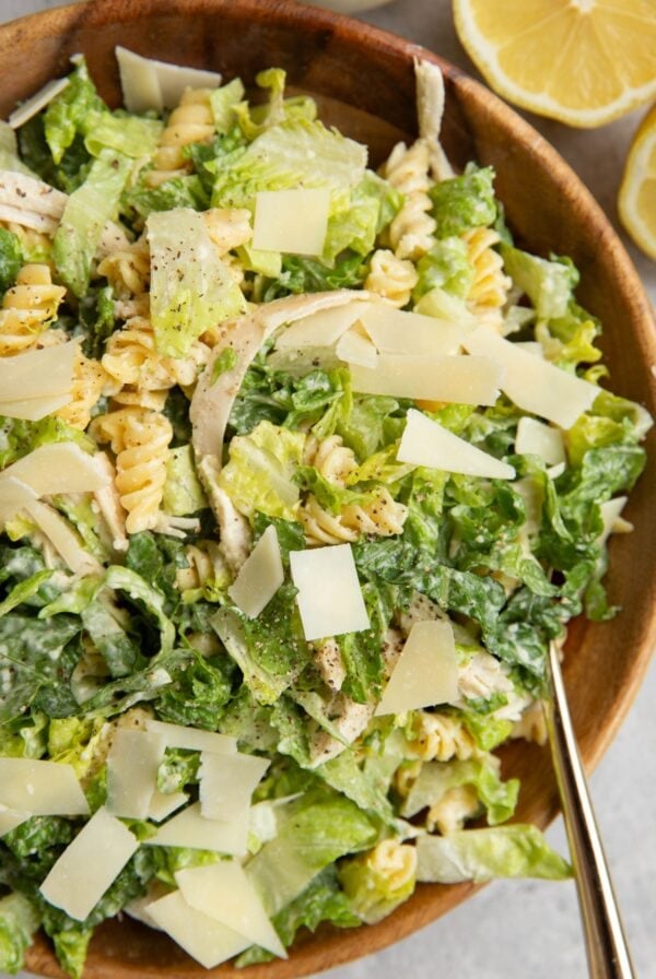 Wooden bowl full of chicken Caesar pasta salad with parmesan cheese on top and a sliced lemon to the side.