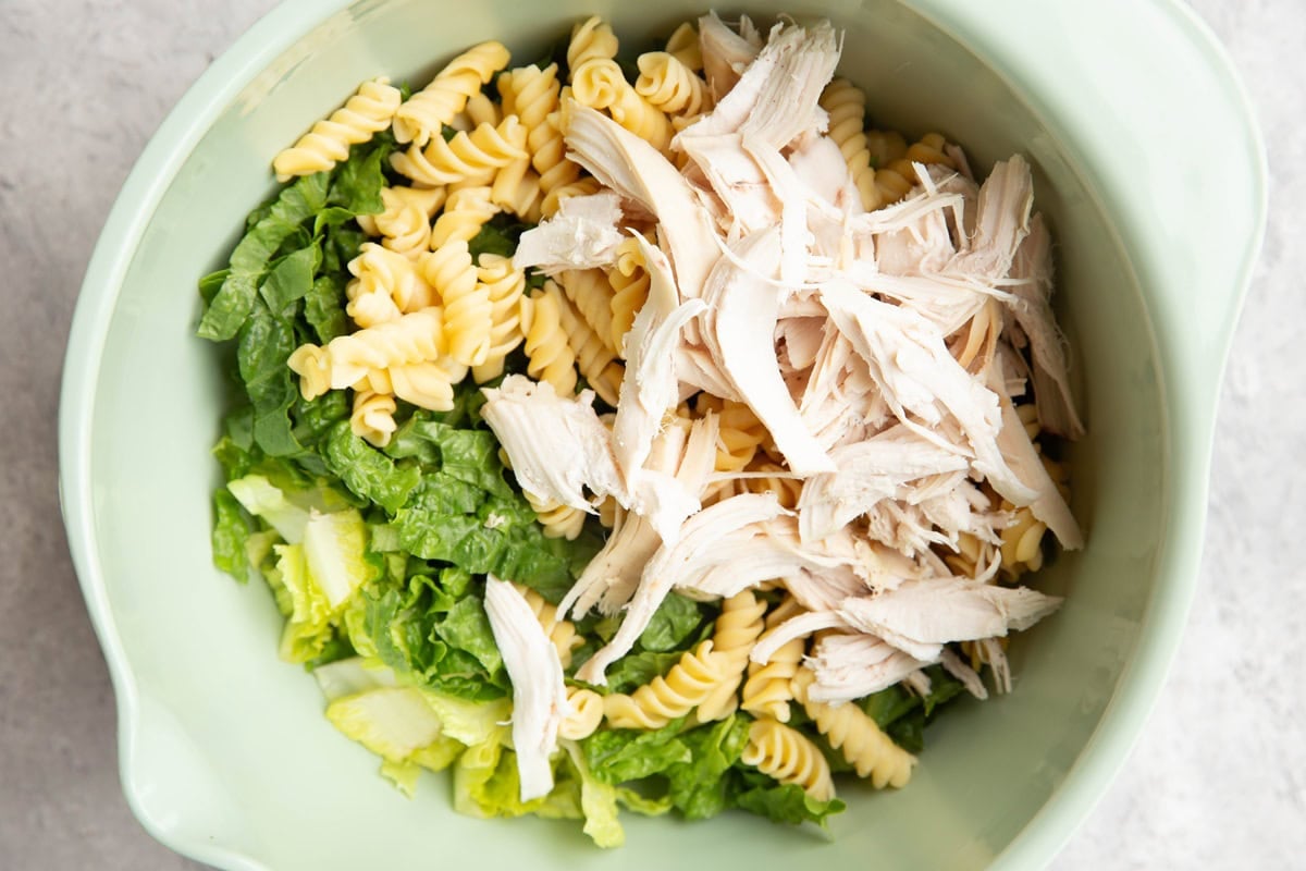 Mixing bowl with chopped lettuce, spiral pasta noodles, and shredded chicken.