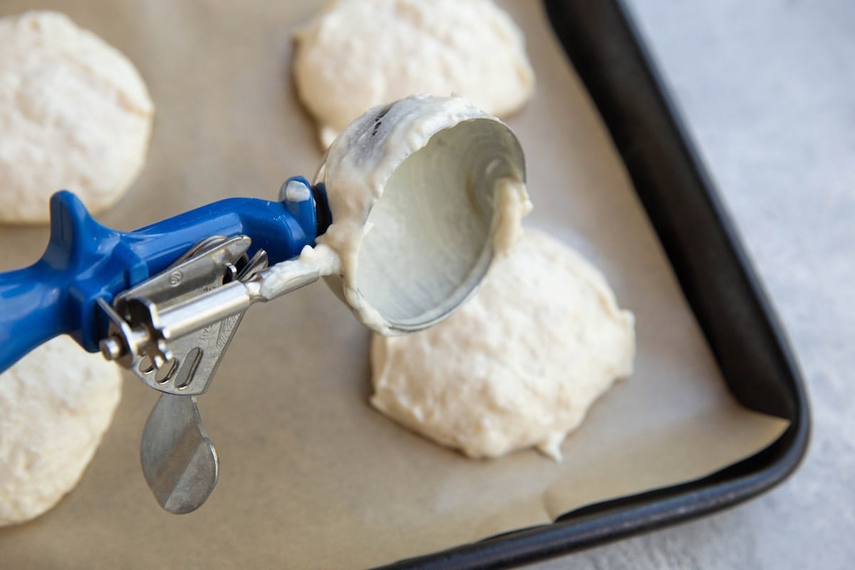 Ice cream scoop scooping mounds of yogurt mixture onto a parchment-lined baking sheet.