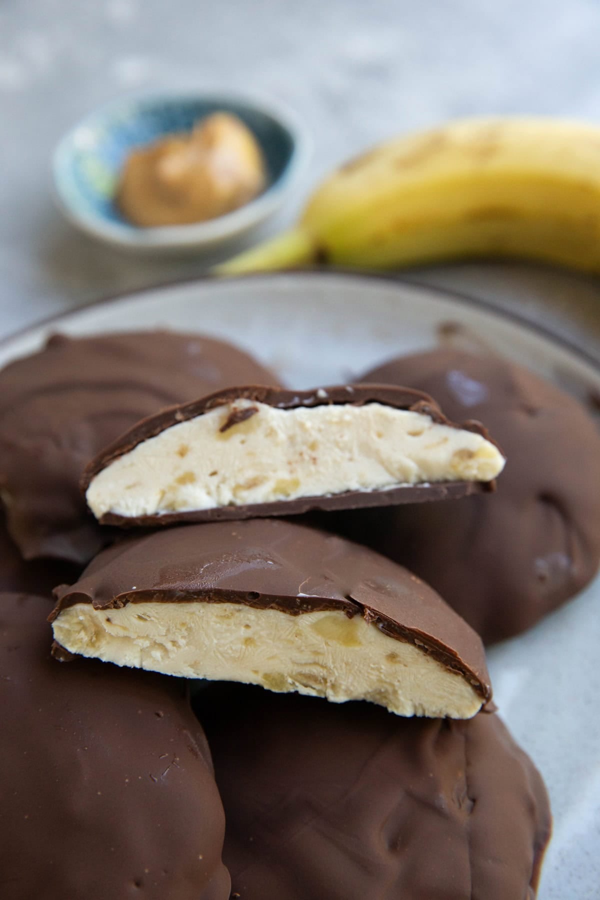Plate with frozen yogurt bite sliced in half to show the frozen yogurt inside. The plate has the rest of the bites with a ripe banana and a bowl of peanut butter in the background.