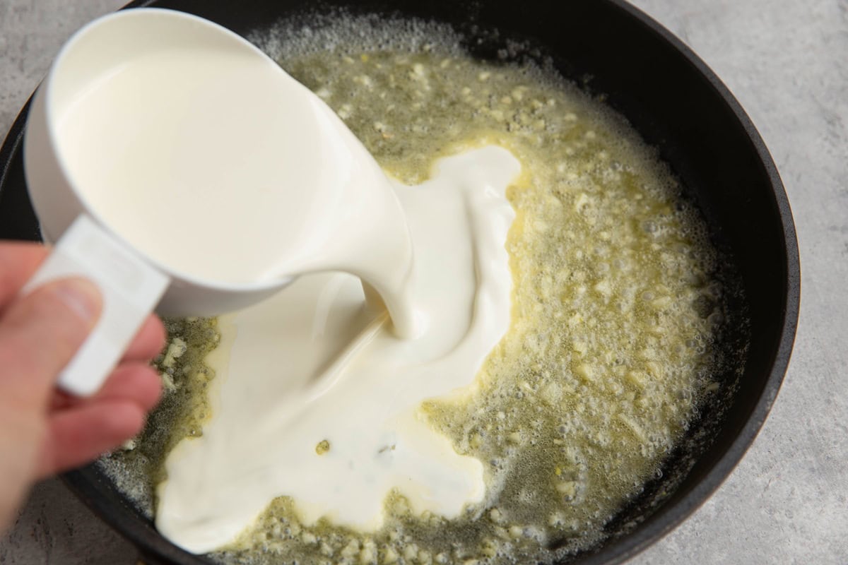 Hand pouring one cup of heavy cream into a skillet with butter and garlic.