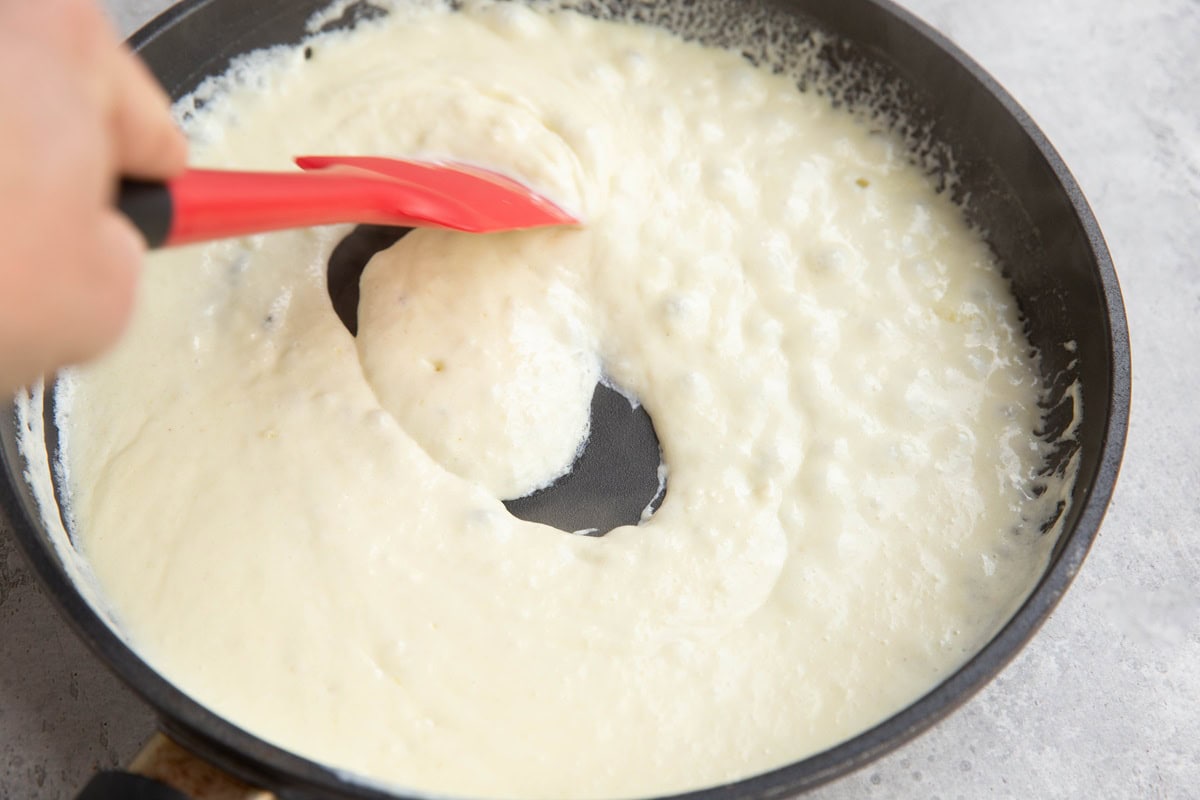White sauce in a skillet cooking at a gentle bubble with a hand stirring the sauce with a rubber spatula.