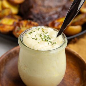 Garlic Butter Sauce in a jar sitting on a wooden plate with a plate of steak and potatoes in the background, ready to be used.