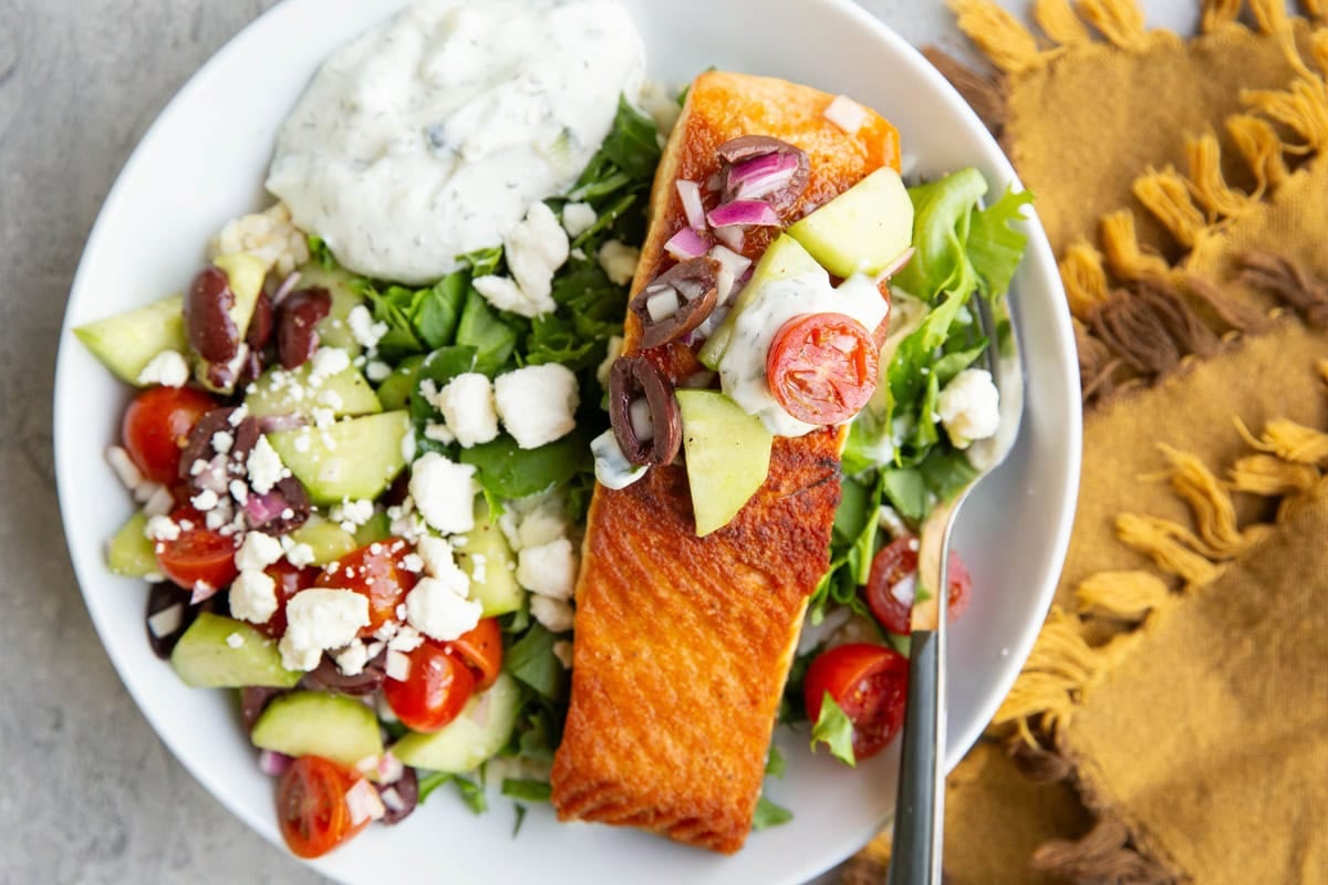 Crispy salmon on a bed of chopped lettuce with cucumber tomato salad, feta cheese and tzatziki sauce in a large bowl with a golden napkin to the side.