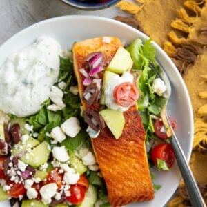 Big white bowl with brown rice, chopped lettuce, crispy seared salmon, Greek salad, feta cheese, and tzatziki sauce with a small blue bowl of more Greek salad to the side and a golden napkin.