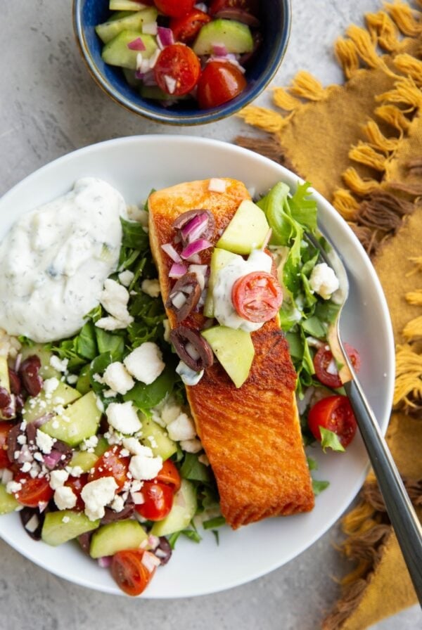 Big white bowl with brown rice, chopped lettuce, crispy seared salmon, Greek salad, feta cheese, and tzatziki sauce with a small blue bowl of more Greek salad to the side and a golden napkin.