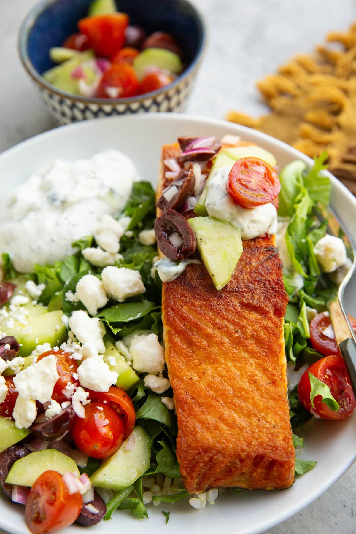 Large white bowl with a close up of crispy salmon topped with cucumber tomato olive salad, feta cheese, and tzatziki sauce. A blue bowl of more vegetables in the background.