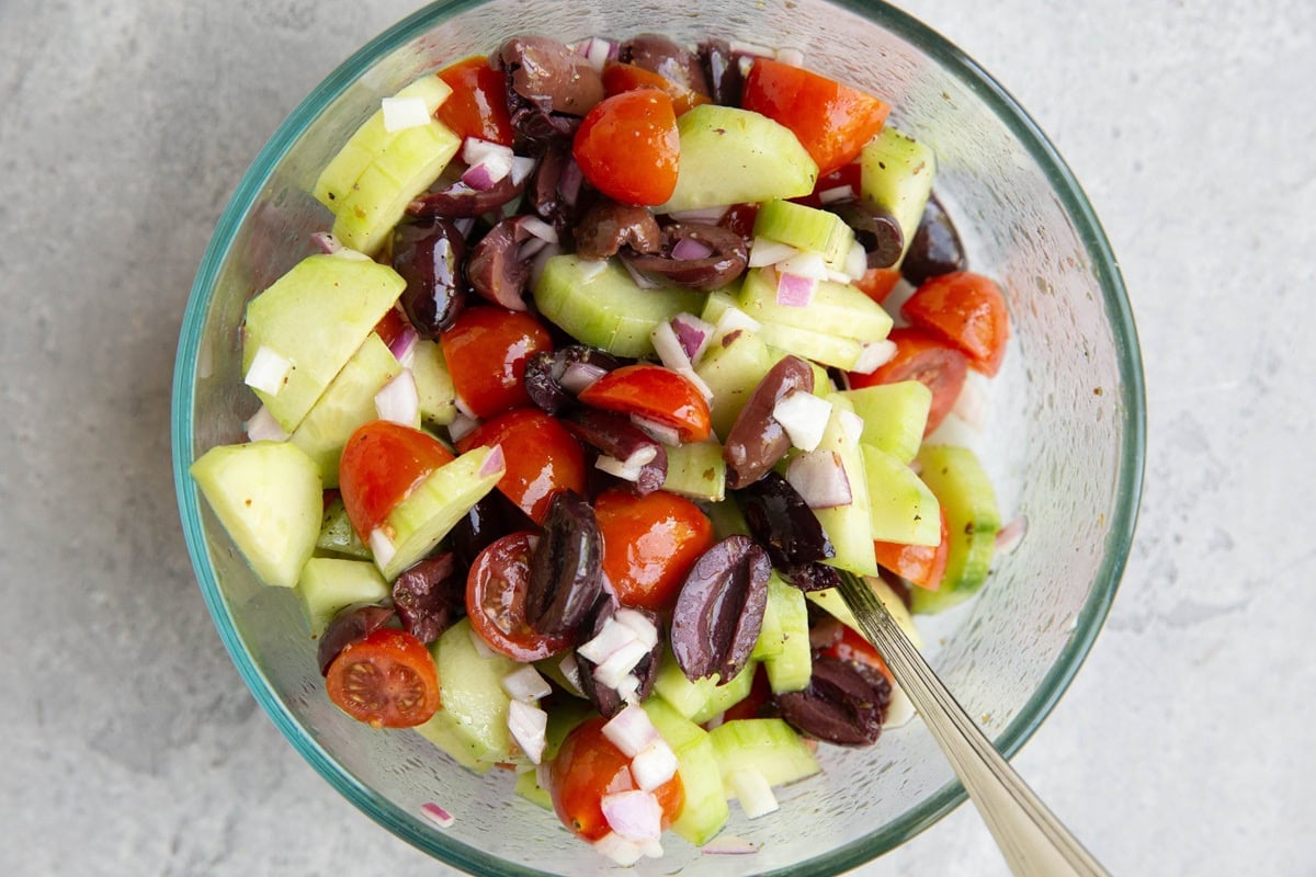 Glass bowl full of chopped cucumbers, cherry tomatoes, kalamata olives, and red onion, all mixed together.