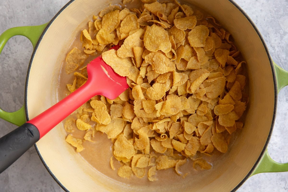A green pot filled with cornflakes being stirred with a red and black silicone spatula, sitting on a light gray surface.