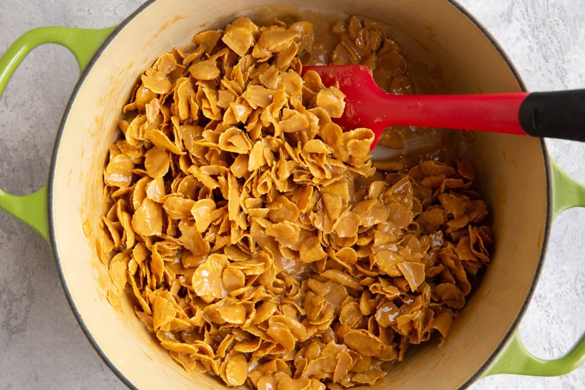 A green Dutch oven pot filled with cornflakes coated peanut butter, honey and butter, being stirred with a red spatula. The pot sits on a light gray countertop.