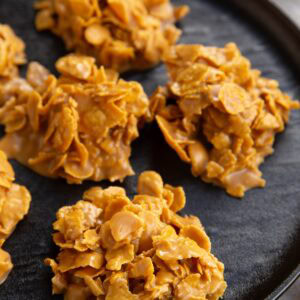 Clusters of golden cornflakes coated in a glossy, peanut butter mixture are arranged on a black plate, resembling homemade no-bake cookies or cereal treats.