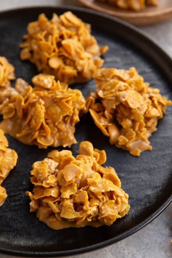 Clusters of golden cornflakes coated in a glossy, peanut butter mixture are arranged on a black plate, resembling homemade no-bake cookies or cereal treats.