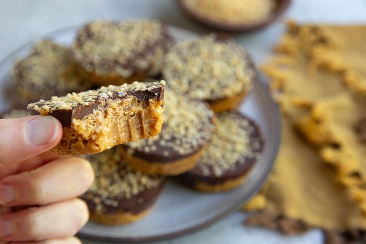 Hand holding a peanut butter cup with a bite taken out and a plate of the rest of the cups in the background.