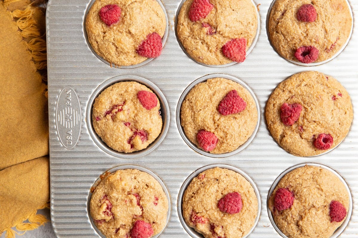 Muffin tray with baked raspberry muffins inside and a golden napkin to the side.