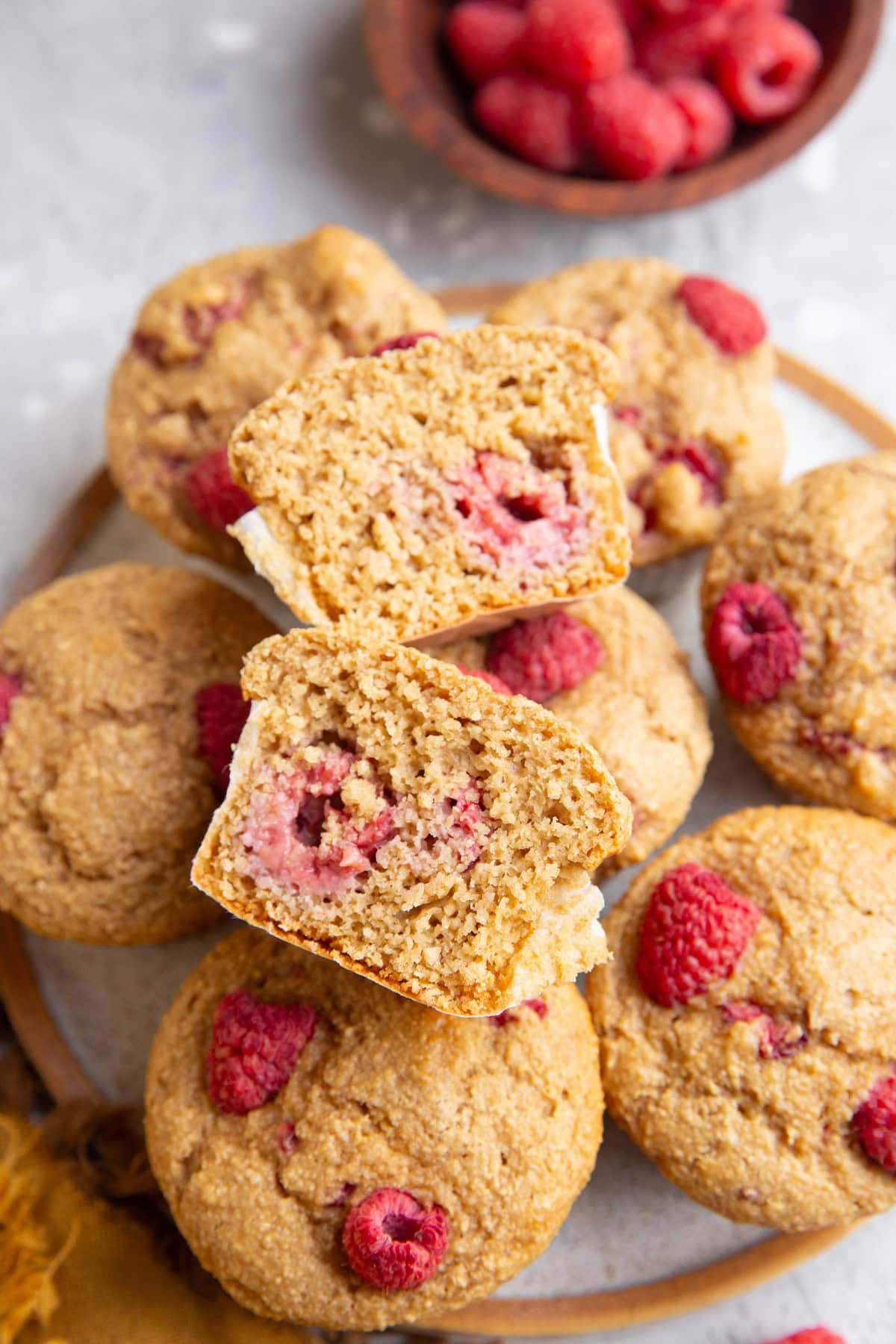 Plate of raspberry muffins with one sliced in half to expose the raspberry inside and a small bowl of fresh raspberries in the background.