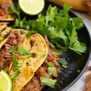 Beef machaca tacos on a black plate with fresh cilantro and limes, ready to serve.