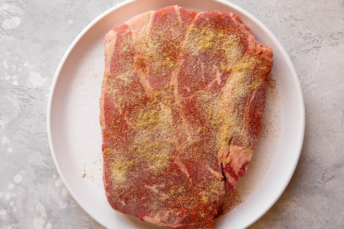 Chuck roast seasoned with paprika, cumin, garlic powder, onion powder and salt on a white plate, ready to be cooked.