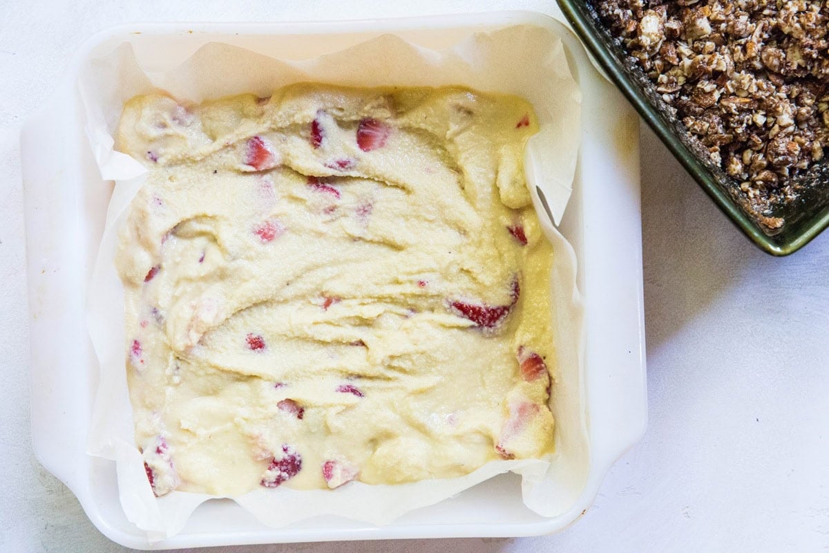 Cake pan lined with parchment paper with strawberry cake batter in an even layer. A bowl of almond topping to the side.