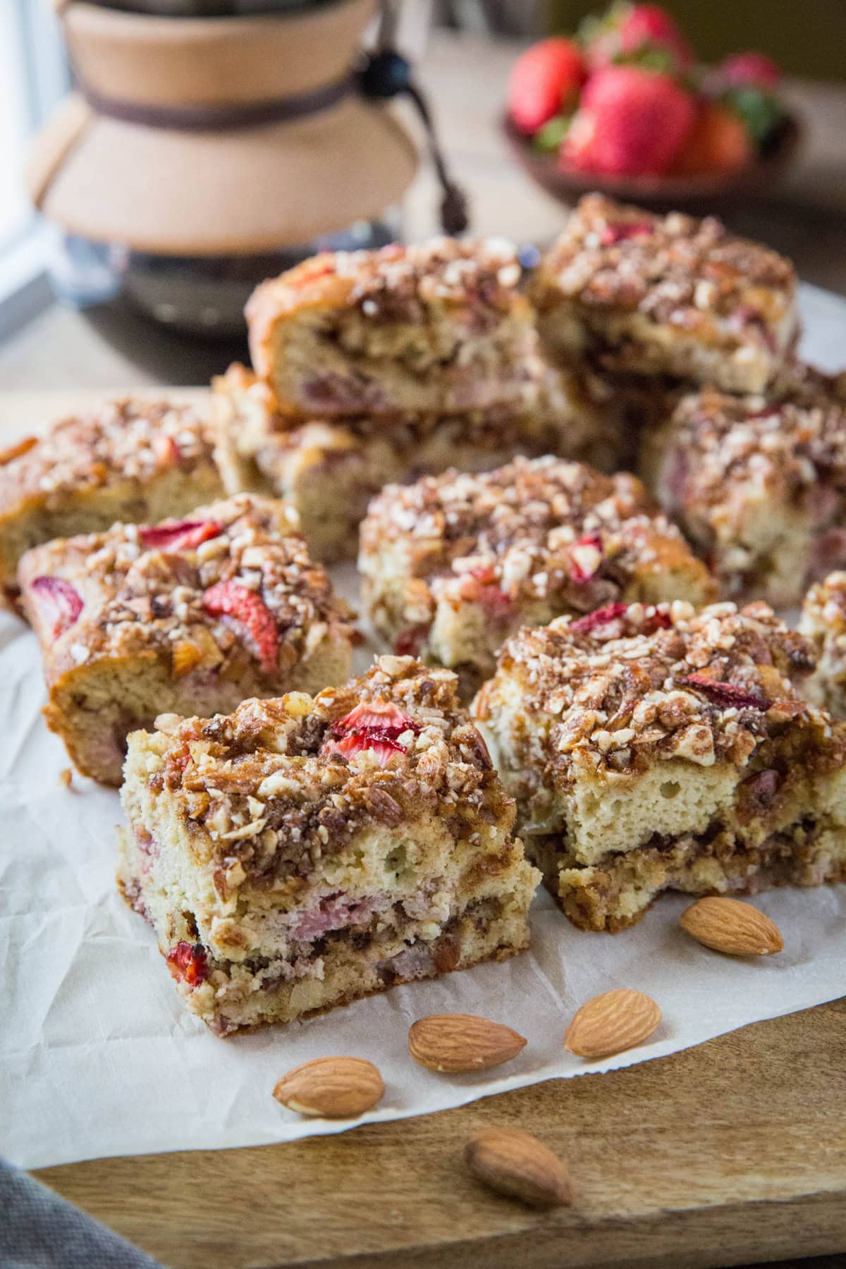 Strawberry crumb cake cut into individual slices on a wooden cutting board with a pour over coffee maker in the background and a small bowl of fresh strawberries.