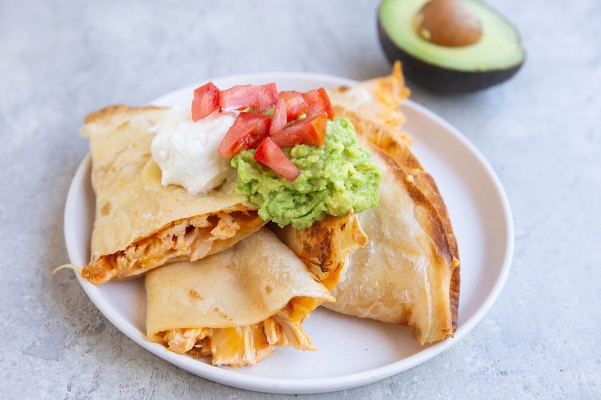 Three chicken quesadillas on a plate with sour cream and guacamole and an avocado in the background.